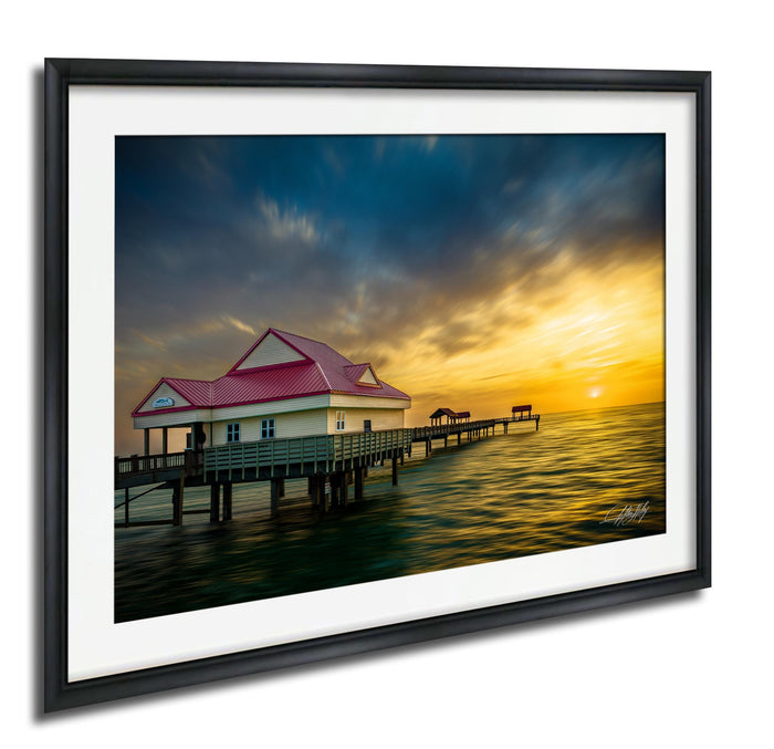 A framed photograph depicting a wooden pier with a red-roofed building, set against a vibrant sunset sky over the ocean.
