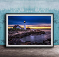 A framed photograph depicting a lighthouse on a rocky shore at sunset, with the sky filled with vibrant colors and the ocean reflecting the warm hues.