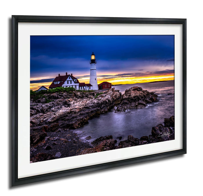 A framed photograph depicting a lighthouse on a rocky shore at sunset, with the sky filled with vibrant colors.