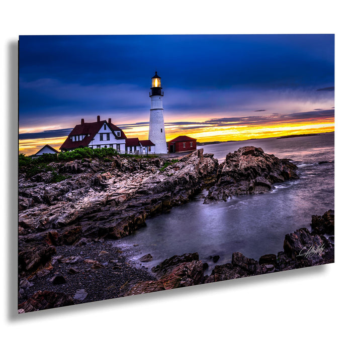 A picturesque coastal scene at sunset, featuring a lighthouse on a rocky shore with a house and a red building in the background. The sky is filled with vibrant colors, creating a stunning backdrop for the lighthouse and the surrounding landscape.
