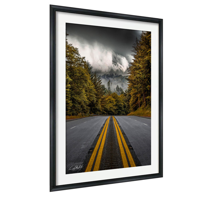 a winding road with yellow lines, surrounded by lush green trees and a dramatic cloudy sky in the background.