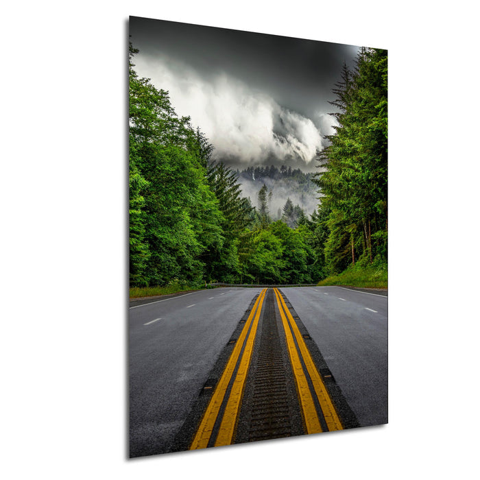 a winding road surrounded by lush green trees and a dramatic cloudy sky in the background.