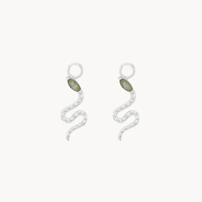 Front view of a pair of silver snake-shaped drop earring charms, each featuring a small marquise green tourmaline gemstone near the head and a textured silver body, set against a plain off-white background.