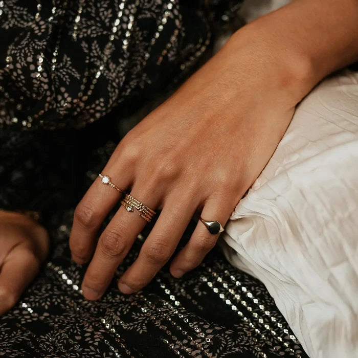 A close-up image of a person's hand adorned with multiple rings, resting on a textured white and black surface. The individual's skin tone is medium, and their fingers are graceful, with one ring featuring small gemstones and another showcasing the revival stacking ring - 14k yellow gold by bluboho.