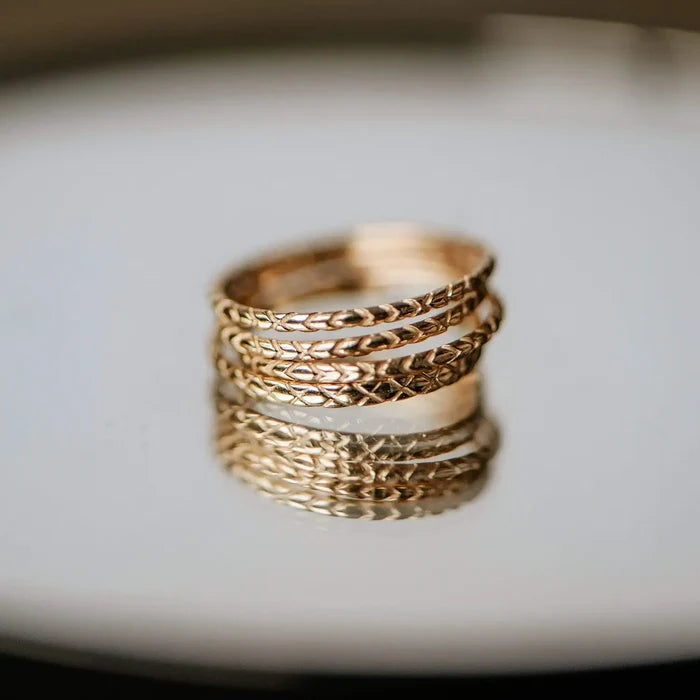 A revival stacking ring - 14k yellow gold featuring three bands intertwined together. Each band has a distinct engraved pattern, with designs including braided and geometric elements. The ring is displayed on a reflective surface, showing a clear reflection of its intricate details by bluboho.