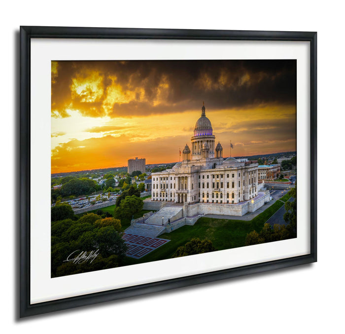 A framed photograph of a large, ornate building with a domed roof, set against a dramatic sky with orange and yellow hues, likely at sunset.
