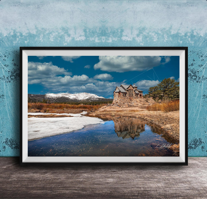 A framed photograph depicting a stone building reflected in a calm body of water, with snow-capped mountains in the background and a cloudy blue sky above.