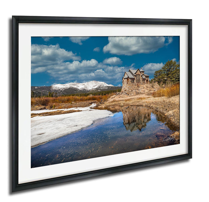 A picturesque landscape with a stone building nestled among snow-capped mountains, surrounded by a serene lake and a clear blue sky with fluffy white clouds.