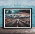 A framed photograph depicting a long, straight road leading towards a snow-capped mountain range under a dramatic, cloudy sky.