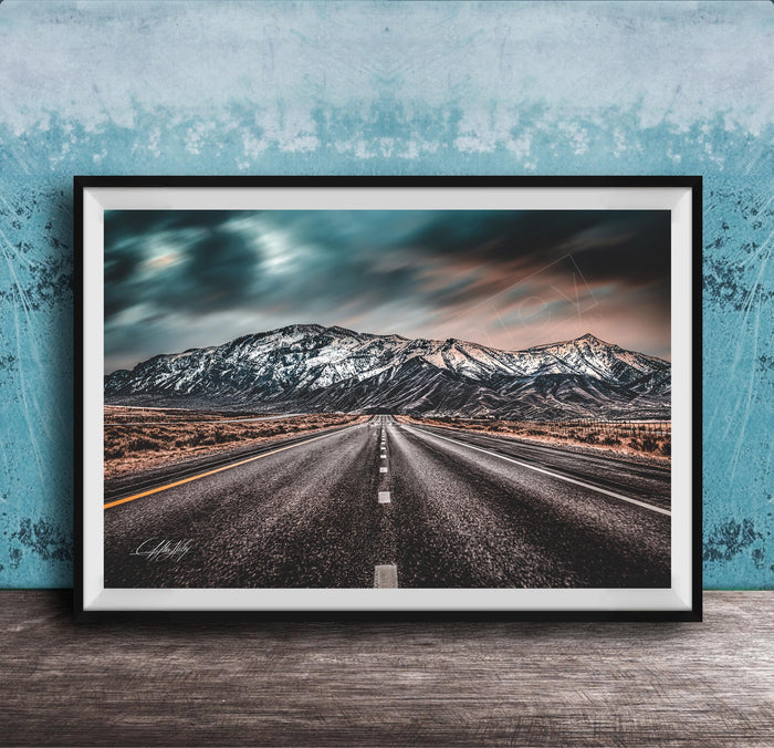 A framed photograph depicting a long, straight road leading towards a snow-capped mountain range under a dramatic, cloudy sky.