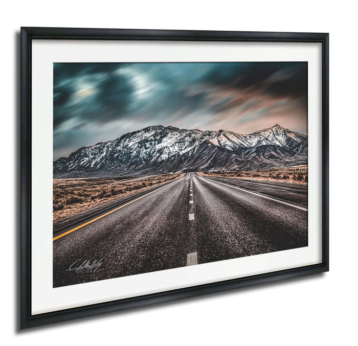 A framed photograph depicting a long, straight road leading towards a snow-capped mountain range under a cloudy sky.