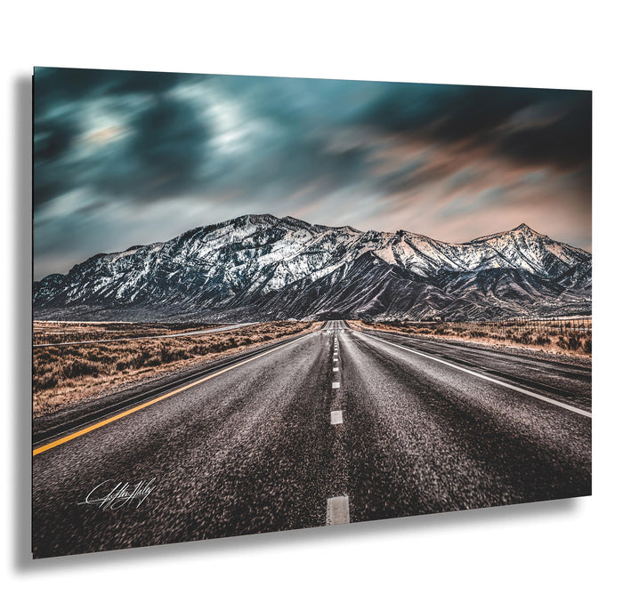A long, straight road stretches out towards a majestic mountain range in the distance, with a cloudy sky overhead.