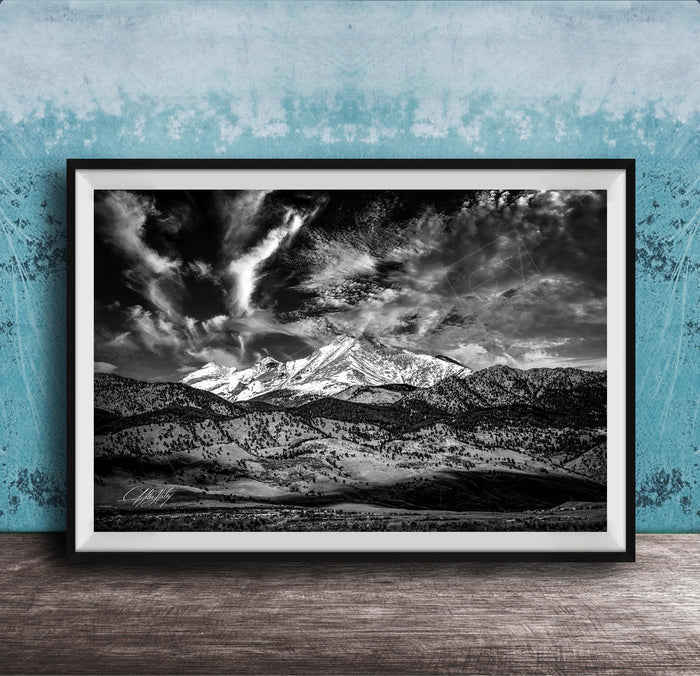 A black and white photograph of a mountainous landscape with snow-capped peaks and dramatic clouds in the sky, framed and displayed on a wooden surface against a blue textured wall.