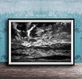 A black and white photograph of a mountainous landscape with snow-capped peaks and dramatic clouds in the sky, framed and displayed on a wooden surface against a blue textured wall.