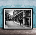 A black and white photograph of a city street lined with buildings and shops, with a cobblestone road and street lamps.
