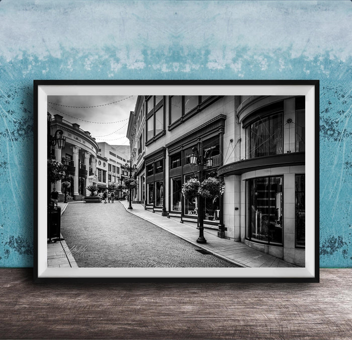 A black and white photograph of a city street lined with buildings and shops, with a cobblestone road and street lamps.
