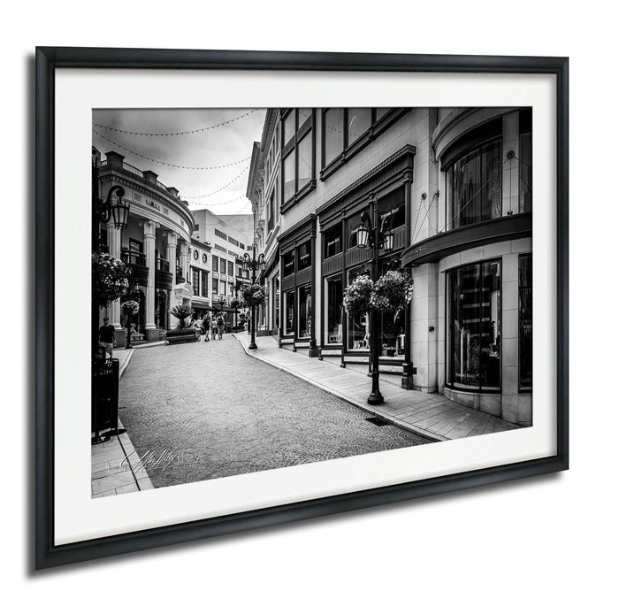 A black and white photograph of a city street lined with buildings, featuring a cobblestone pavement and street lamps.