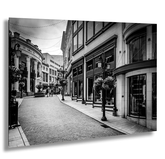 A black and white photograph depicting a city street lined with buildings, shops, and people walking along the sidewalk.