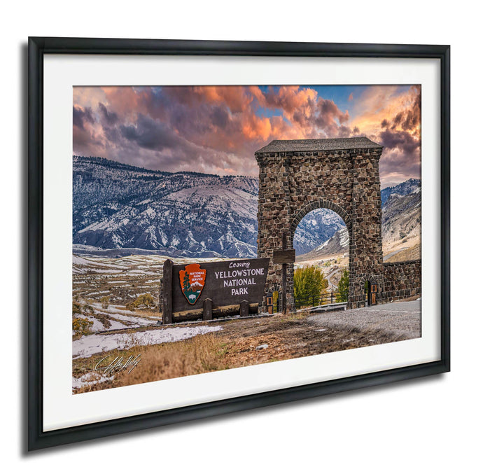 A framed photograph of the iconic Yellowstone National Park arch, with a scenic mountain landscape in the background and a colorful sunset sky.