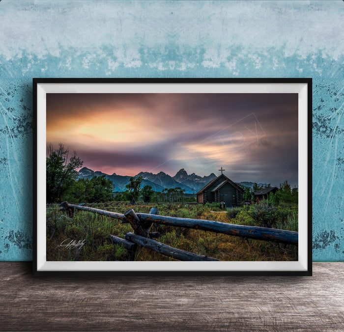 A framed photograph depicting a serene rural landscape with a wooden fence, a church, and mountains in the distance, set against a colorful sunset sky.