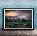 A framed photograph depicting a serene rural landscape with a wooden fence, a church, and mountains in the distance, set against a colorful sunset sky.