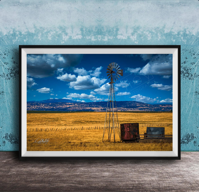 A framed photograph depicting a windmill standing in a field with a blue sky and clouds in the background.
