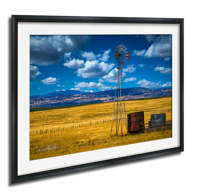 A windmill stands tall in a vast, golden field under a blue sky dotted with fluffy white clouds.