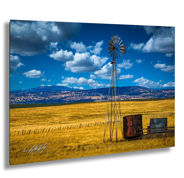 A windmill stands tall in a vast, golden field under a blue sky dotted with fluffy white clouds.