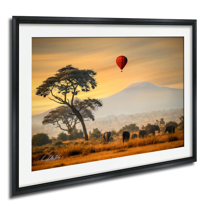 A framed photograph depicting a serene landscape with a lone tree, a herd of elephants, and a hot air balloon in the sky, all set against a warm, golden sunset.
