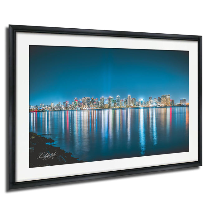 A framed photograph depicting a city skyline at night, with the buildings' lights reflected in the water below.