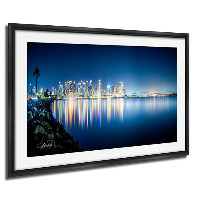 A framed photograph depicting a city skyline at night, with the city's lights reflected in the water below.