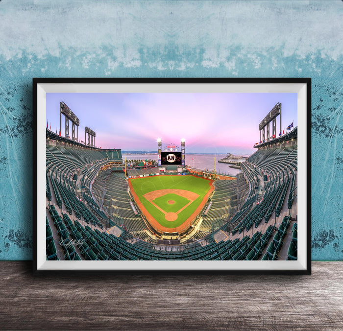 A framed photograph of a baseball stadium at dusk, with the field illuminated and the sky displaying a colorful sunset.