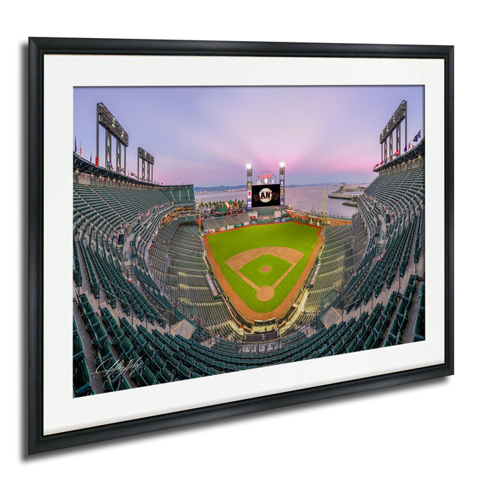 A framed photograph of a baseball stadium with a large scoreboard and field, set against a colorful sunset sky.