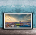 The image is a framed photograph of a cityscape at sunset, featuring a skyline with tall buildings and a body of water in the foreground.