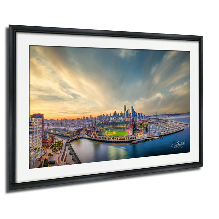 The image is a framed aerial view of a city skyline at sunset, with the city's buildings and landmarks illuminated against a colorful sky.
