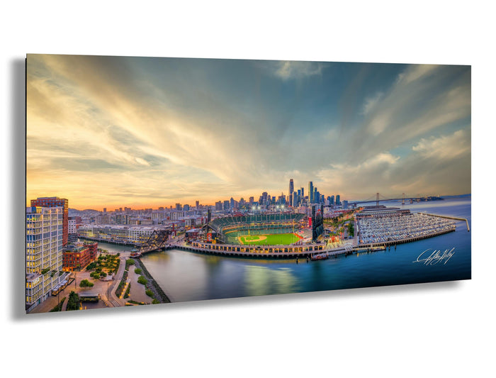 A panoramic view of a city skyline at sunset, with a baseball field and a body of water in the foreground.