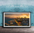 The image is a framed photograph of a cityscape at sunset, featuring a skyline with tall buildings and a harbor with boats.