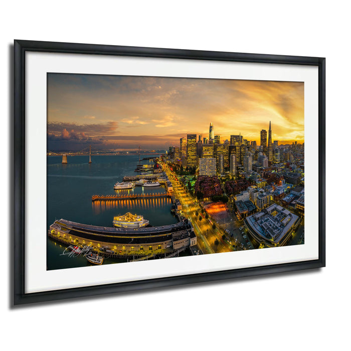 The image is a framed aerial view of a city skyline at sunset, with the city lights illuminating the buildings and the water below.