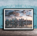 A framed photograph of a city skyline at sunset, with the buildings reflected in the water below.