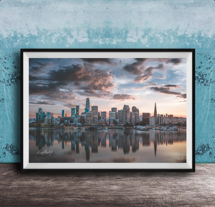 A framed photograph of a city skyline at sunset, with the buildings reflected in the water below.