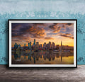 A framed photograph depicting a city skyline at sunset, with the city's buildings reflected in the water below.