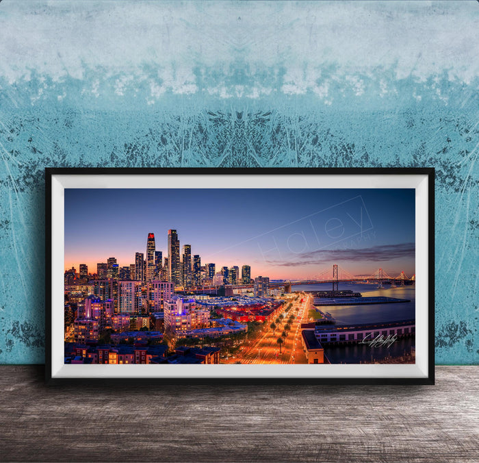A framed photograph of a city skyline at dusk, with the city lights illuminating the buildings and the water in the background.