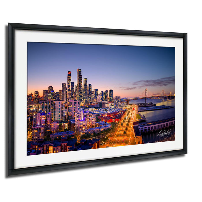 The image is a framed photograph of a city skyline at dusk, with the city lights illuminating the buildings and the surrounding area.