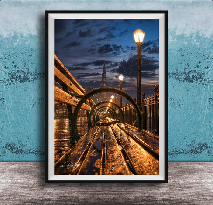 A wooden bench on a bridge at night, with a city skyline and street lamps in the background, creating a serene and atmospheric scene.