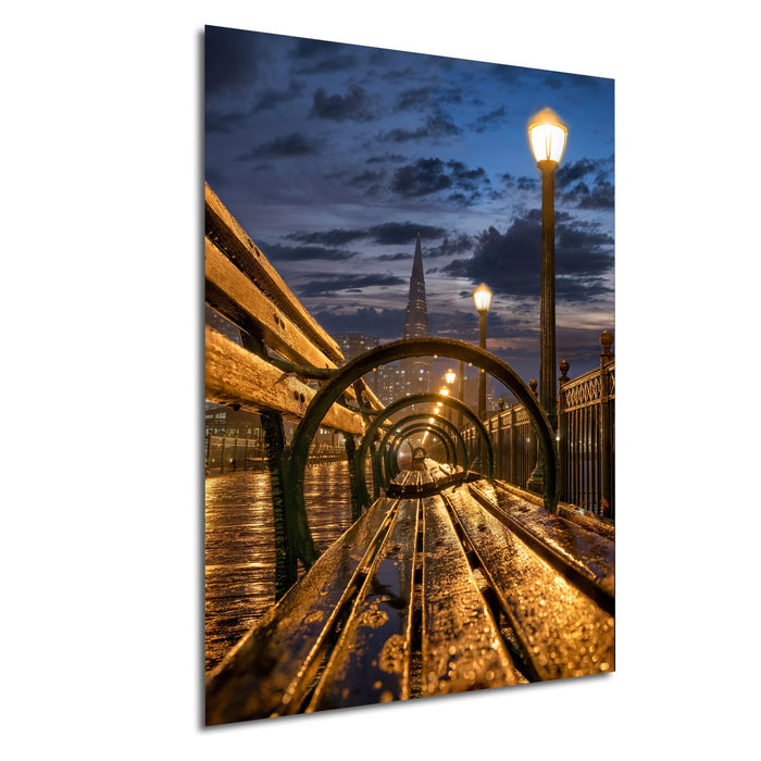 A wooden bench sits on a wet, reflective surface, with a curved metal railing and street lamps in the background, creating a serene and atmospheric nighttime scene.