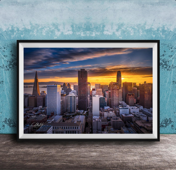 The image is a framed photograph of a city skyline at sunset, with the sun setting behind the buildings and casting a warm, golden glow over the urban landscape.