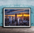 The image is a framed photograph of a city skyline at sunset, with the sun setting behind the buildings and casting a warm, golden glow over the urban landscape.