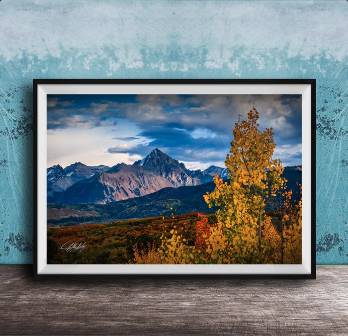 The image is a framed photograph depicting a scenic mountain landscape with a mix of autumnal and winter colors, including vibrant yellow and orange foliage in the foreground and majestic snow-capped peaks in the background.