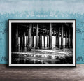 A black and white photograph of a pier with multiple columns and pillars, set against a blue textured wall.