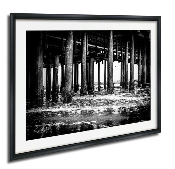 A black and white photograph of a pier with numerous wooden columns and beams, partially submerged in water.